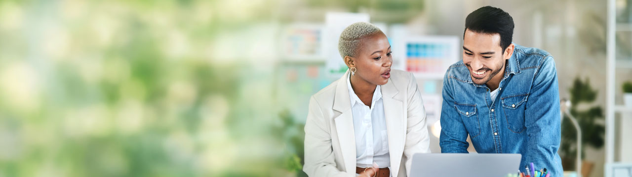 Mujer con blazer beige y hombre con camisa vaquera mirando un portátil, con plantas al fondo